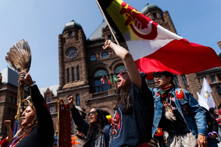 A low-angle shot of protestors, some with fists raised, one holding a flag, with the Ontario Legislature visible in the background.