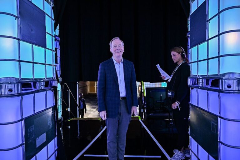 Brad Smith, vice-chair and president of Microsoft, stands in a hallway backstage at Web Summit Vancouver 2025 at the Vancouver Convention Centre, flanked by glowing blue screens.