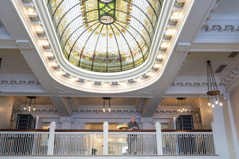 A person stands on a balcony inside a building with a large stained glass dome ceiling and ornate architectural details.
