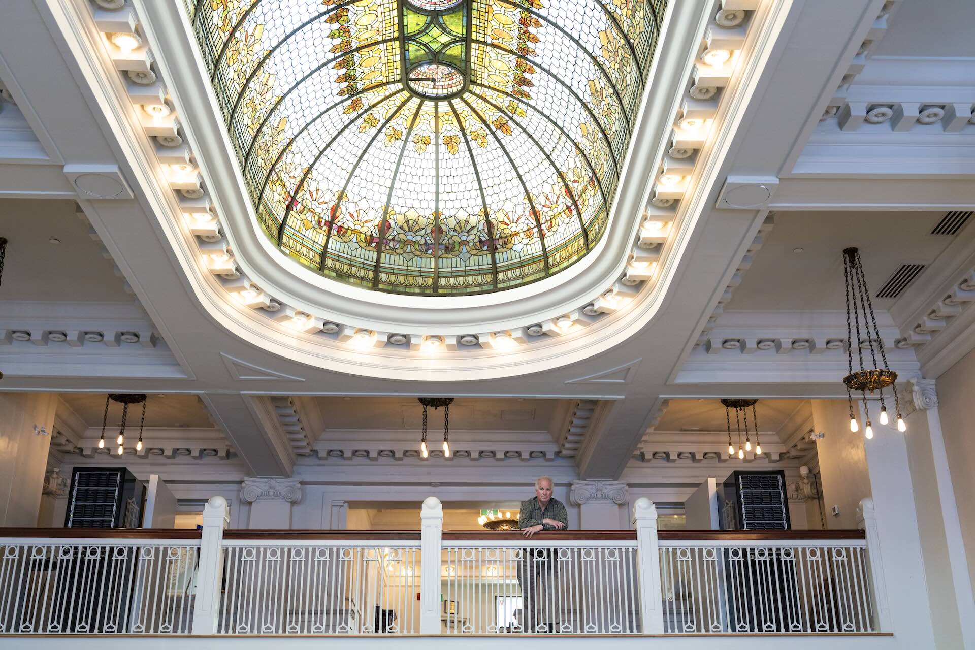 A person stands on a balcony inside a building with a large stained glass dome ceiling and ornate architectural details.