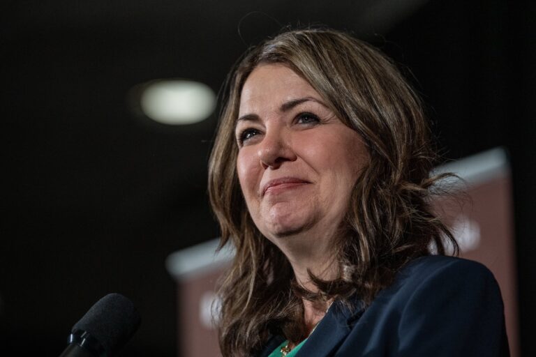 A close-up of Danielle Smith in front of a microphone. She's wearing an arch facial expression, with a close-lipped smile.