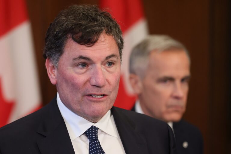 A close-up of Dominic LeBlanc at a news conference. Prime Minister Mark Carney is visible, out of focus, over LeBlanc's left shoulder.
