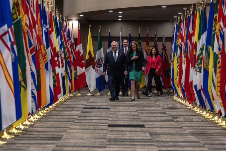 A head-on shot of Doug Ford and Danielle Smith walking toward the camera down an aisle lined by provincial and Canadian flags.
