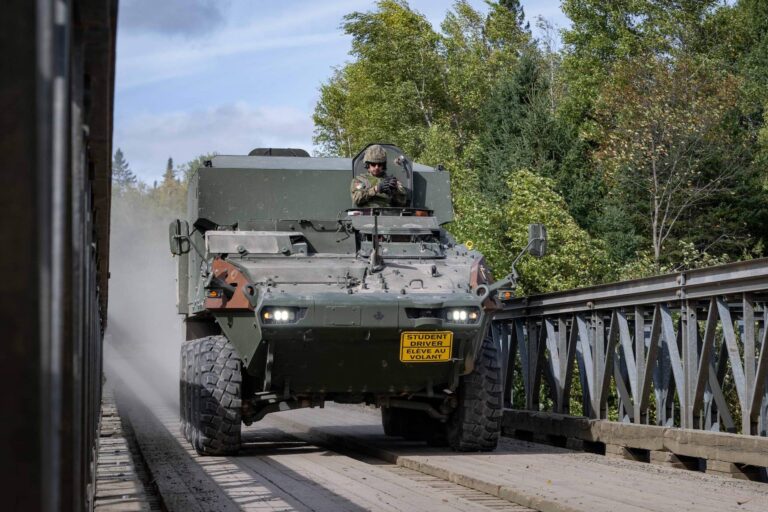 A head-on shot of an eight-wheeled military vehicle crossing a bridge, kicking up dust in its wake. A helmeted soldier is sitting in the top hatch near the front.