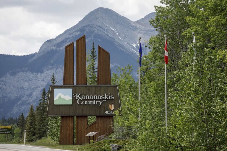 A shot of the sign welcoming highway travellers to Kananaskis Country in Alberta. There are leafy trees in the foreground and mountains looming in the background.