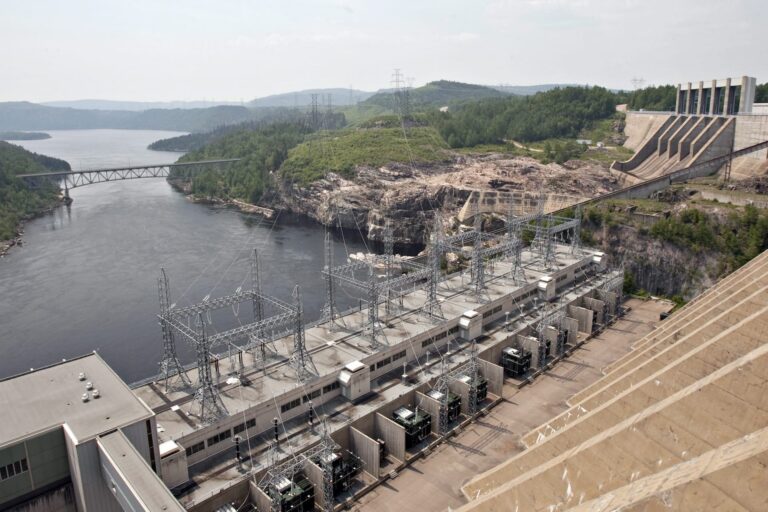 A large hydroelectric dam with transmission lines spans a river, with forested hills, a bridge, and industrial structures visible in the background.