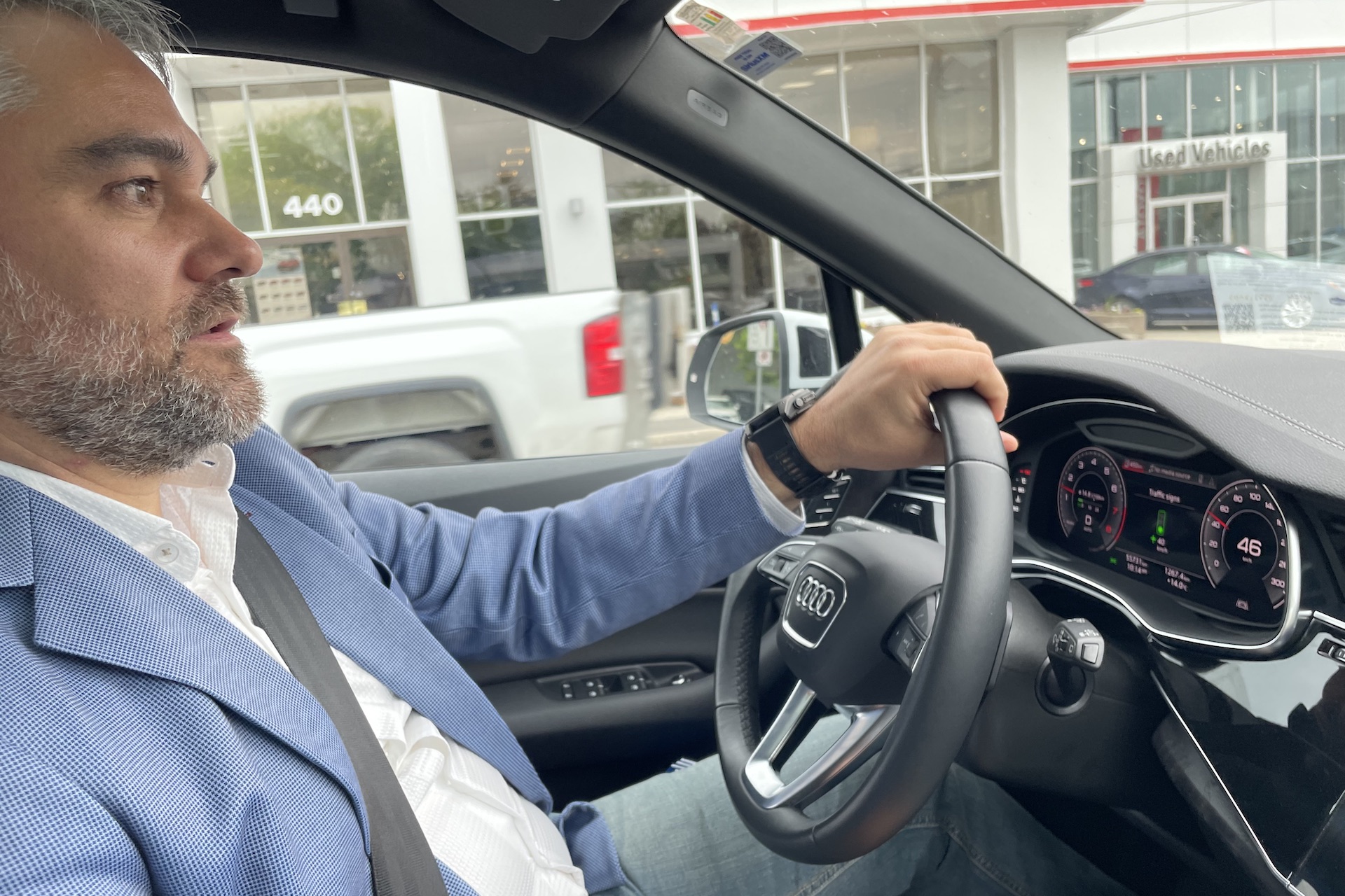 A side shot of Kurtis McBride behind the wheel of an Audi SUV. He's wearing a light blue blazer and jeans. The dashboard display reads "46," and shows an icon of a green traffic light.