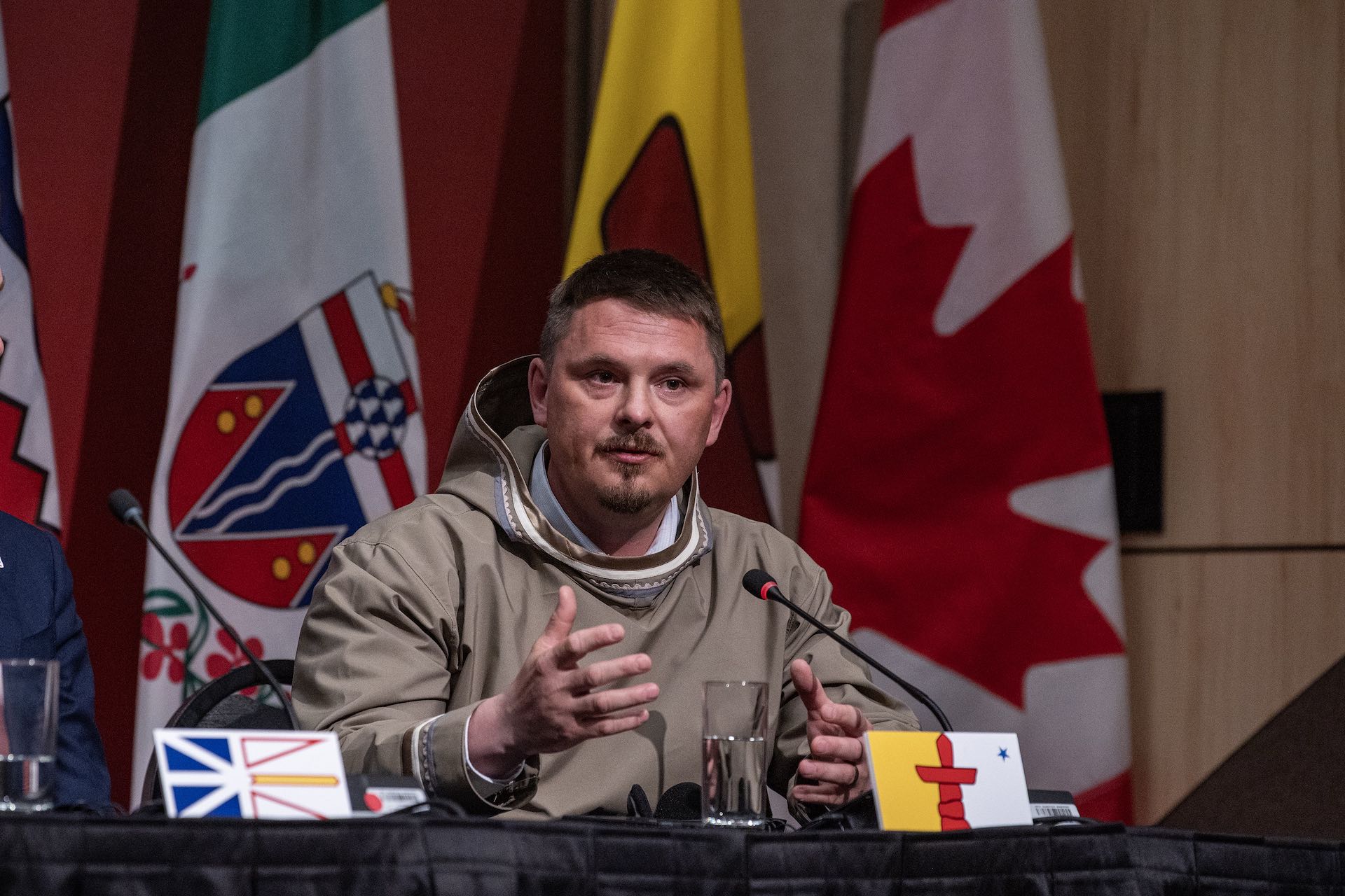 A shot of P.J. Akeeagok at a press conference table with a small placard showing the Nunavut flag in front of him. He's wearing a beige hooded pullover with what appears to be traditional patterning on the cuffs and hood.