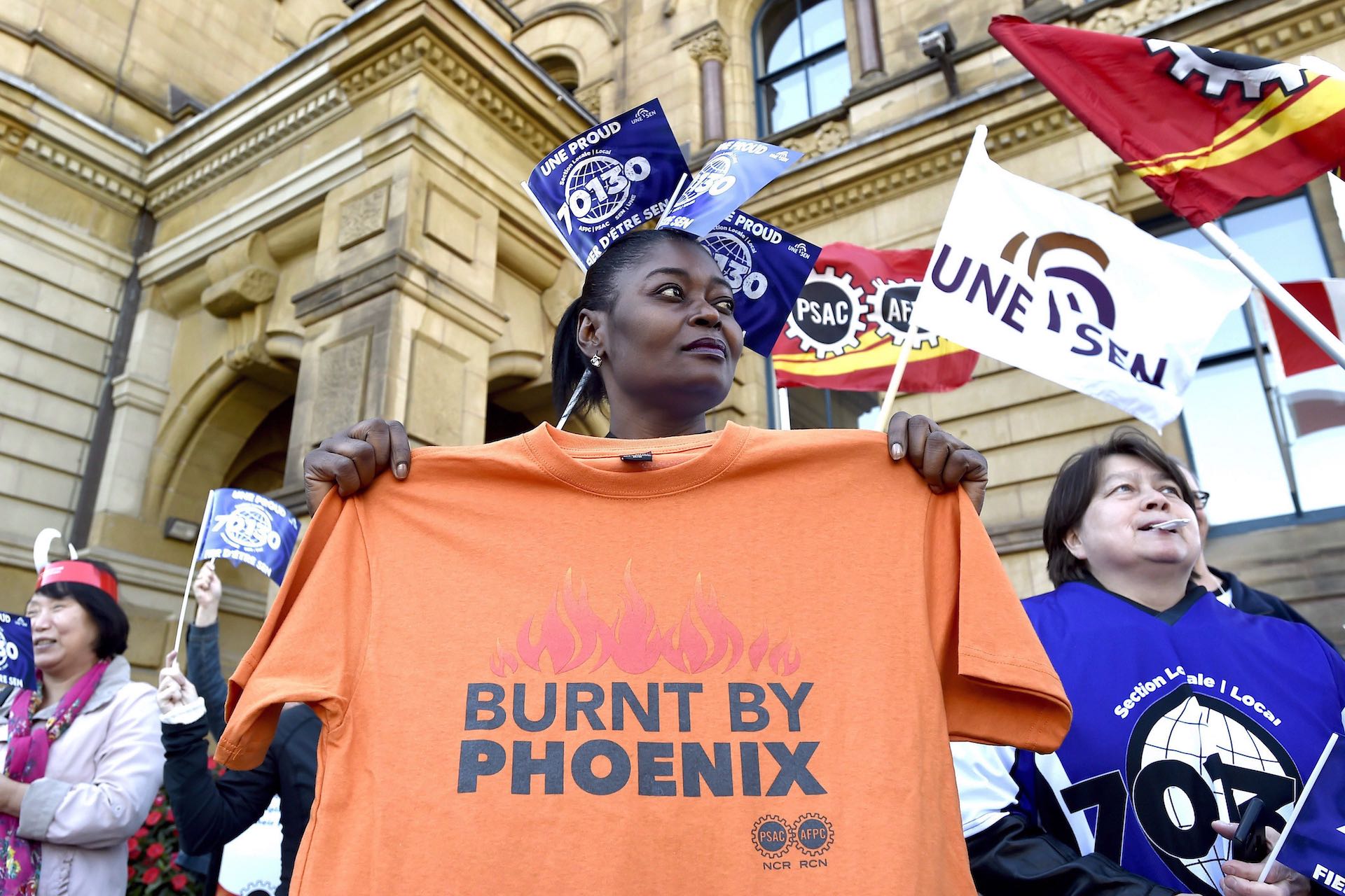 A low-angle shot of a woman holding an orange t-shirt that reads "Burnt by Phoenix." She is standing amid fellow protesters, some of whom are waving flags bearing union insignia.