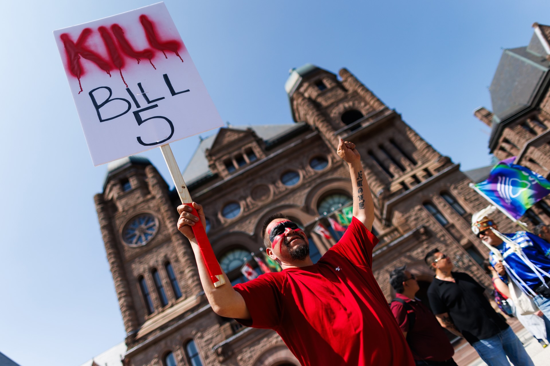 A low-angle shot of an Indigenous protester holding up a sign that reads "Kill Bill 5." He's wearing a red shirt and face paint, and the Ontario legislative building is looming in the background.