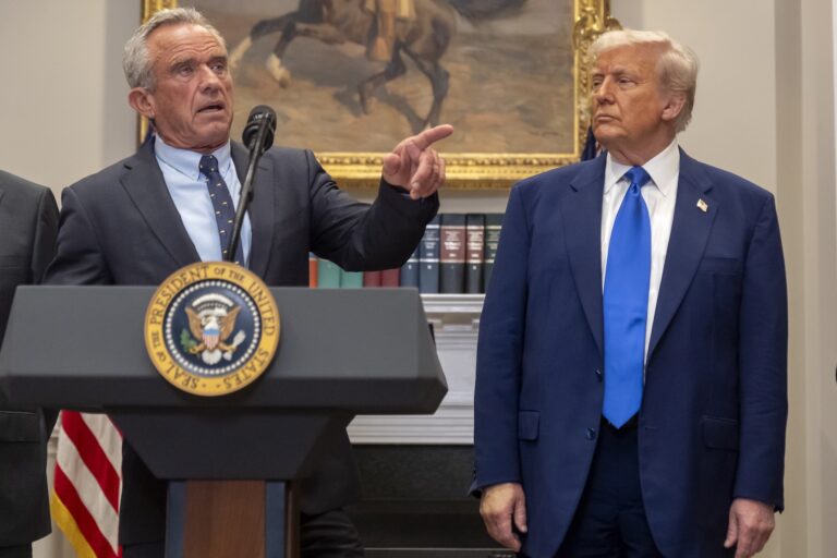 A lower-angle shot of Robert F. Kennedy Jr. behind a lectern with the U.S. presidential seal on the front. He is pointing off-camera. Donald Trump is standing to Kennedy's left.
