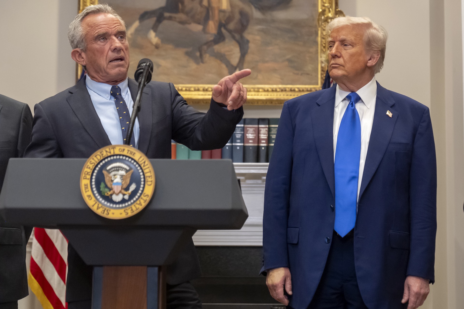A lower-angle shot of Robert F. Kennedy Jr. behind a lectern with the U.S. presidential seal on the front. He is pointing off-camera. Donald Trump is standing to Kennedy's left.