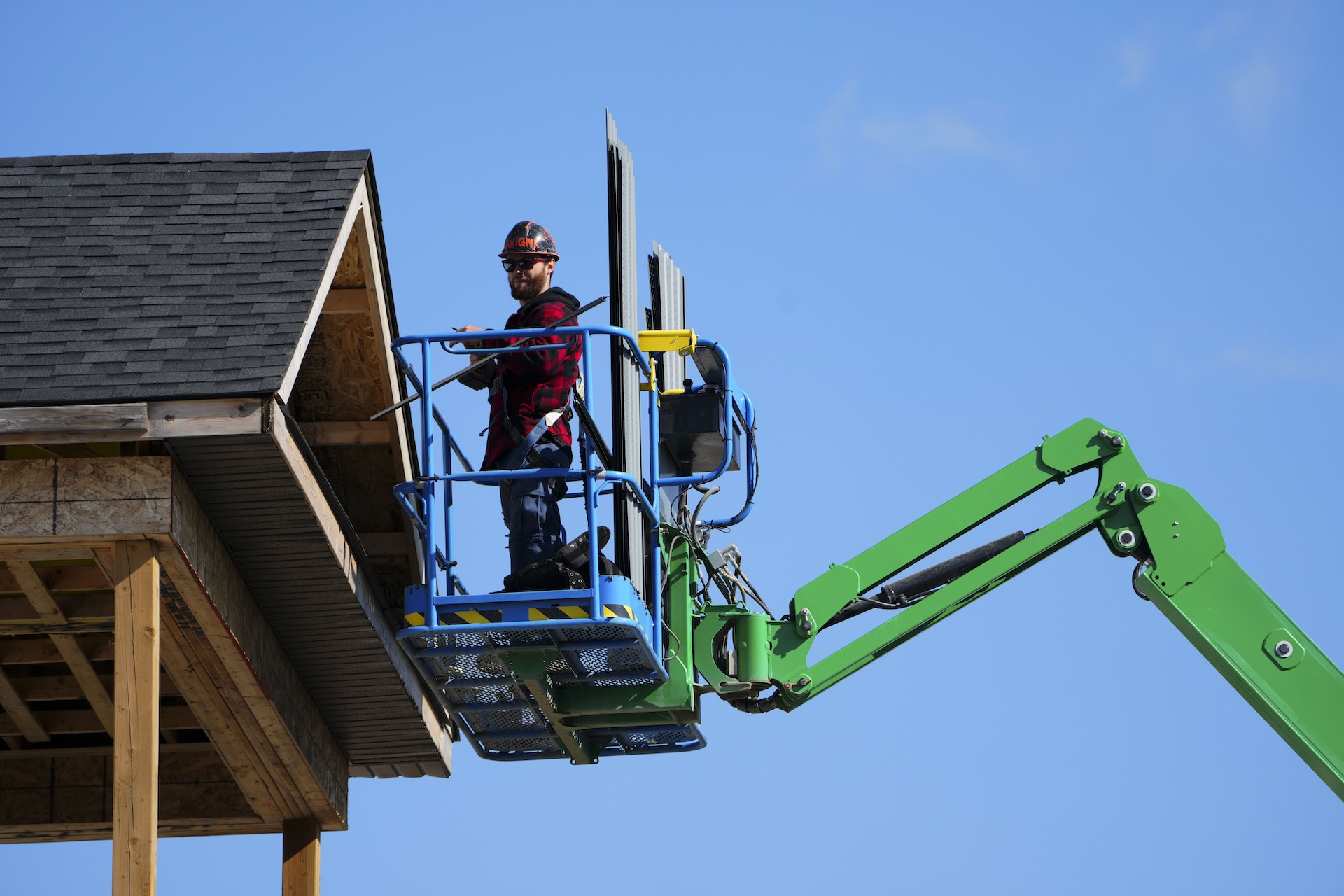 A construction worker stands on a green boom lift, working on the exterior of a building under a clear blue sky.