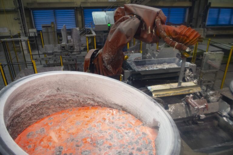 A robotic arm operates near a large container filled with molten aluminum inside an industrial facility.