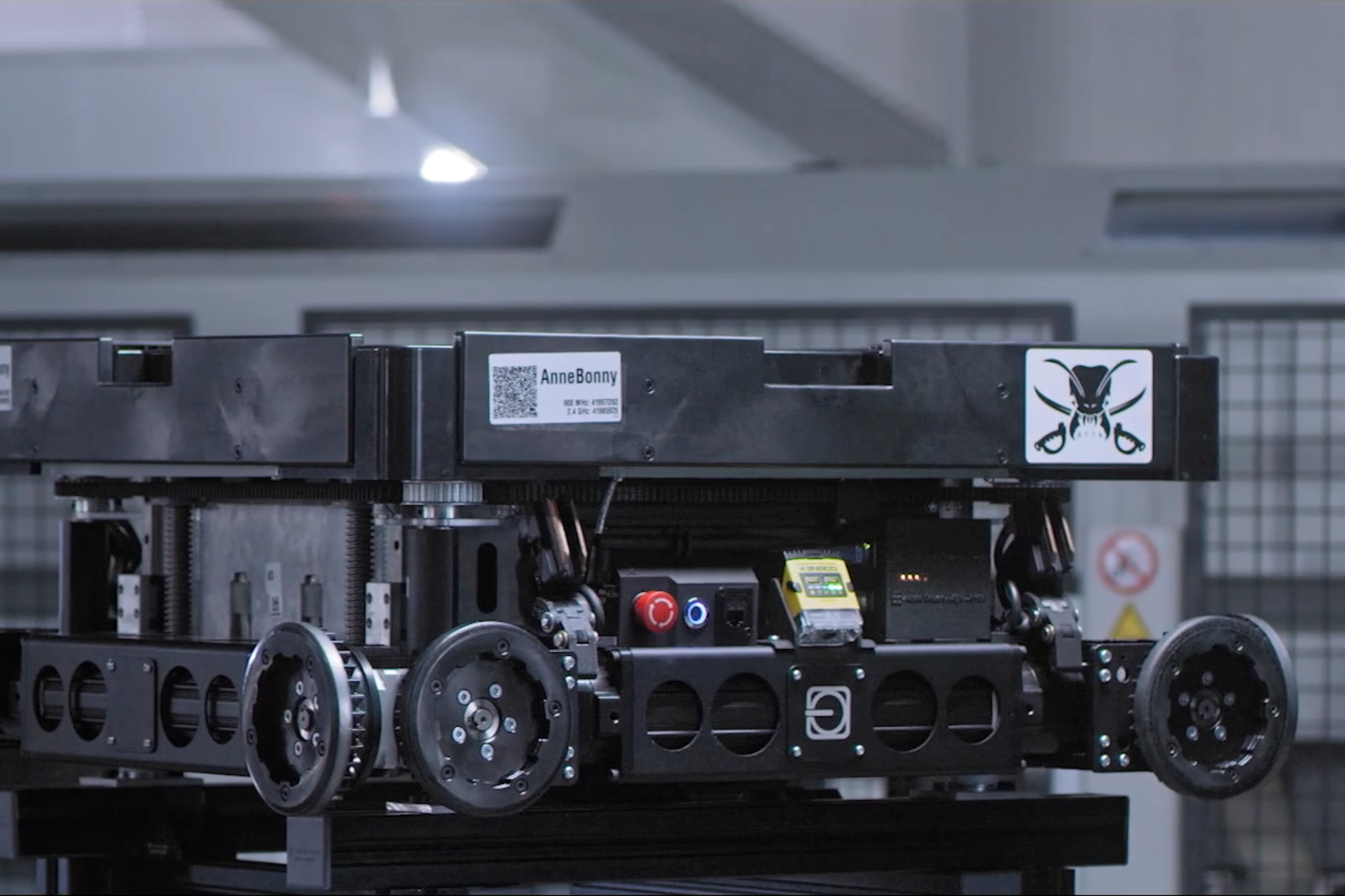 A close-up of a black rectangular robotic unit with wheel-like devices on the bottom. There is a black-and silver logo affixed to the side of it featuring an ant's head and crossed swords.