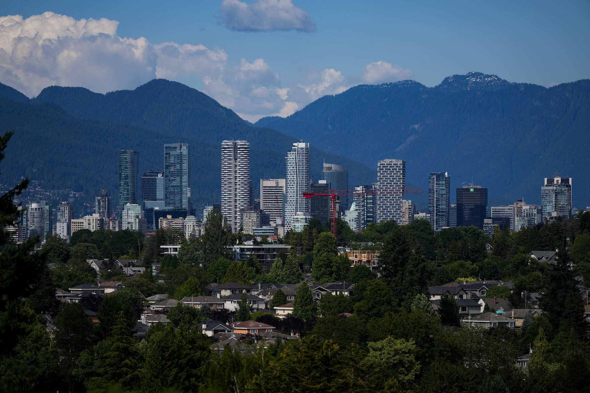 The Vancouver city skyline with high-rise buildings and construction cranes, set against a backdrop of forested mountains and partly cloudy skies.