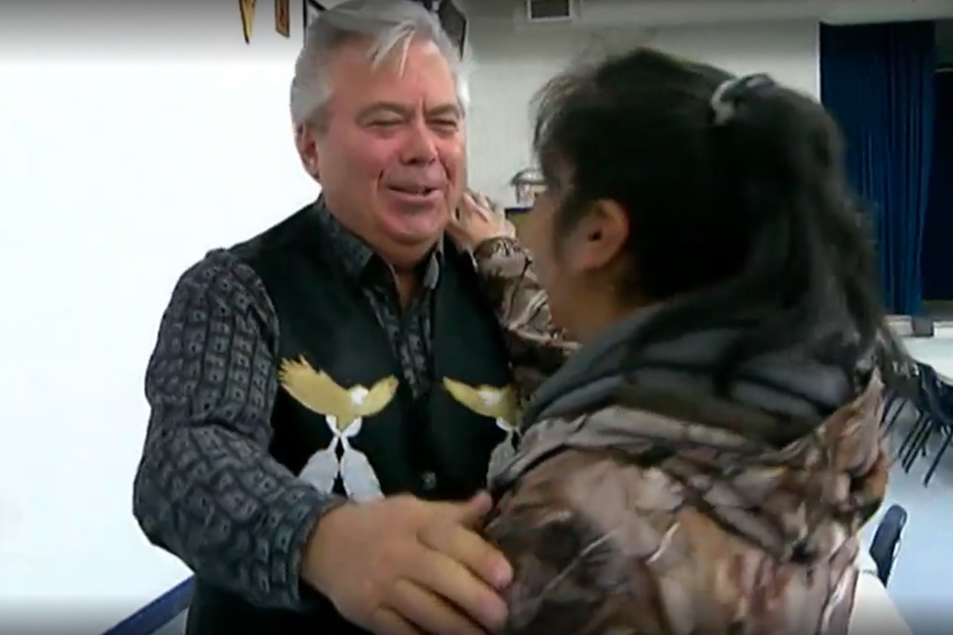 A shot of a smiling, red-faced Darryl Williams who is wearing a black vest with images if an eagle and feathers. It is taken over the shoulder of a woman wearing a camouflage-pattered jacket, who is in the process of embracing Williams.