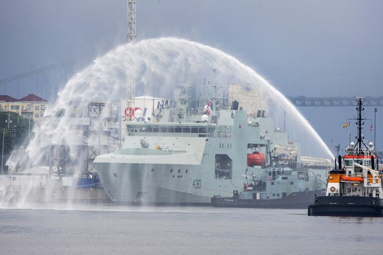 A shot of a grey naval patrol ship passing beneath a celebratory arch of water being sprayed from an adjacent tugboat.