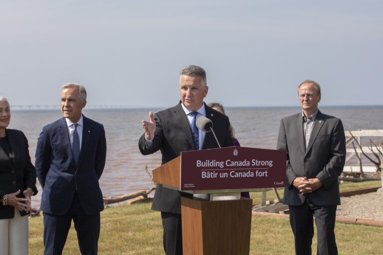 A man speaks at a podium labeled "Building Canada Strong" outdoors by the water, with three people standing nearby.