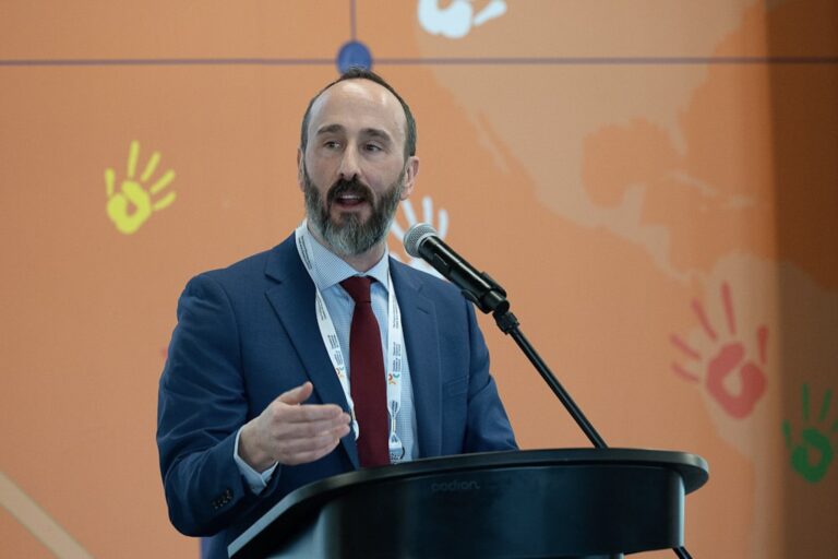 A head-and-shoulders shot of Jay Barber speaking behind a microphone and podium. He has a beard, and is wearing a blue jacket with a burgundy tie. There is an orange backdrop behind him.