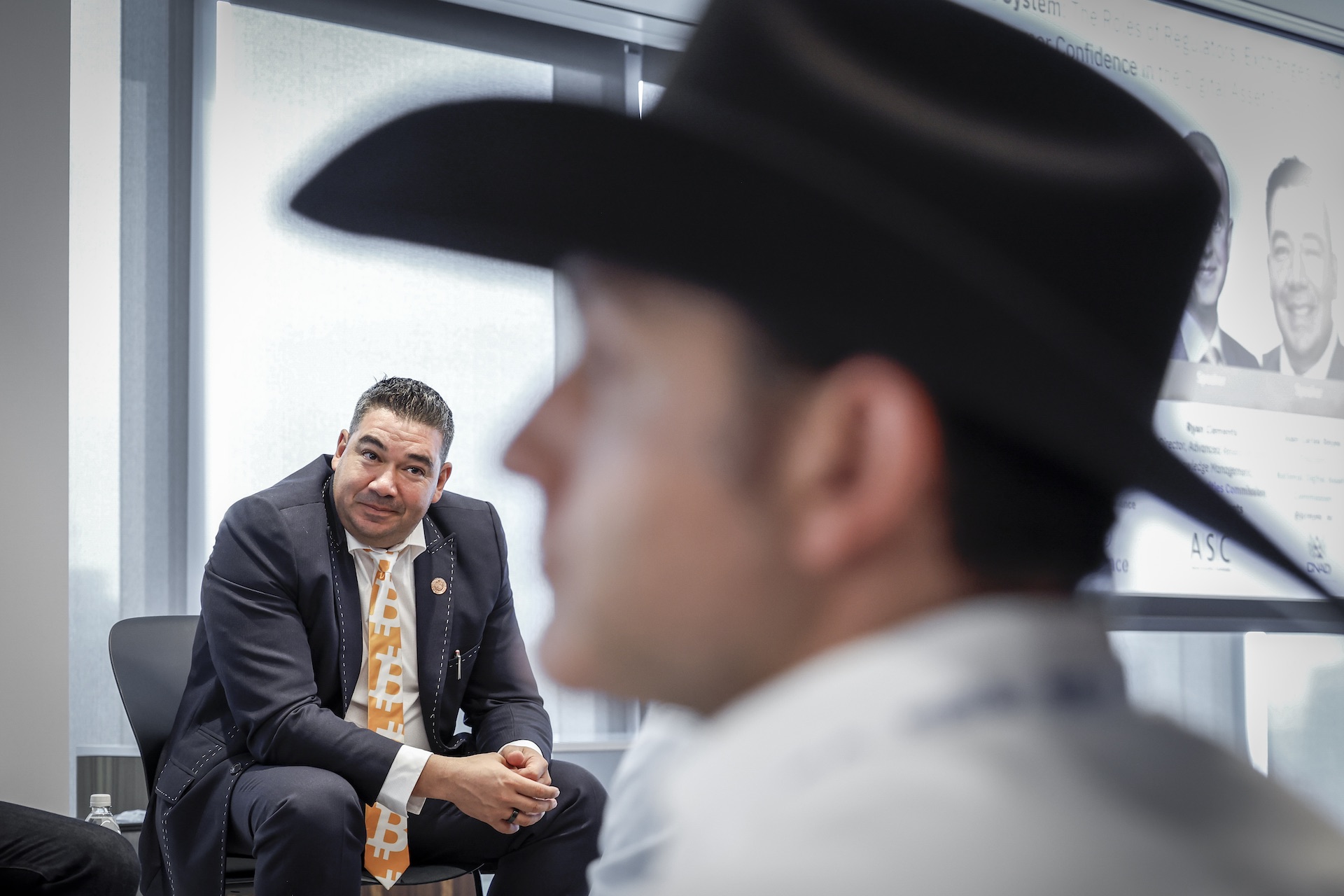 A shot of Juan Carlos Reyes seated on a stage, leaning forward with his hands folded. There is a man wearing a black cowboy hat out of focus in the foreground.