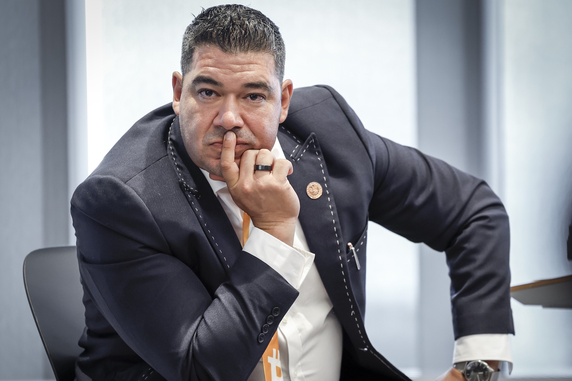 A face-on shot of Juan Carlos Reyes seated on-stage at an event. He is looking at directly and the camera with his elbow on his knee and chin in his hand. He's wearing a dark jacket with white outer stitching and an orange tie that features the Bitcoin logo.