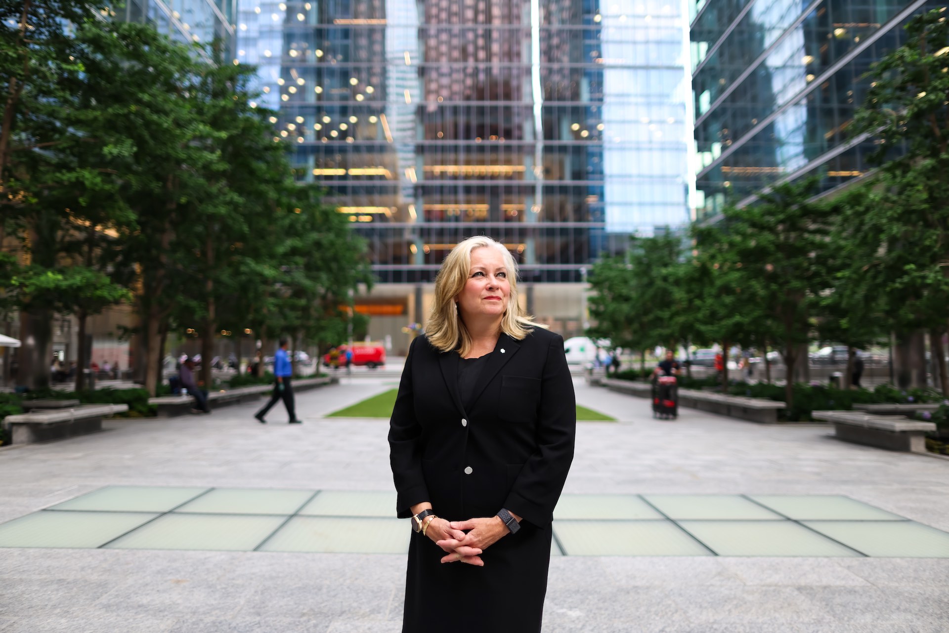 A portrait-style shot of Linda Hasenfratz in an open city square, with trees and glass office towers in the background. She is wearing a black dress jacket and has her hands clasped in front of her.