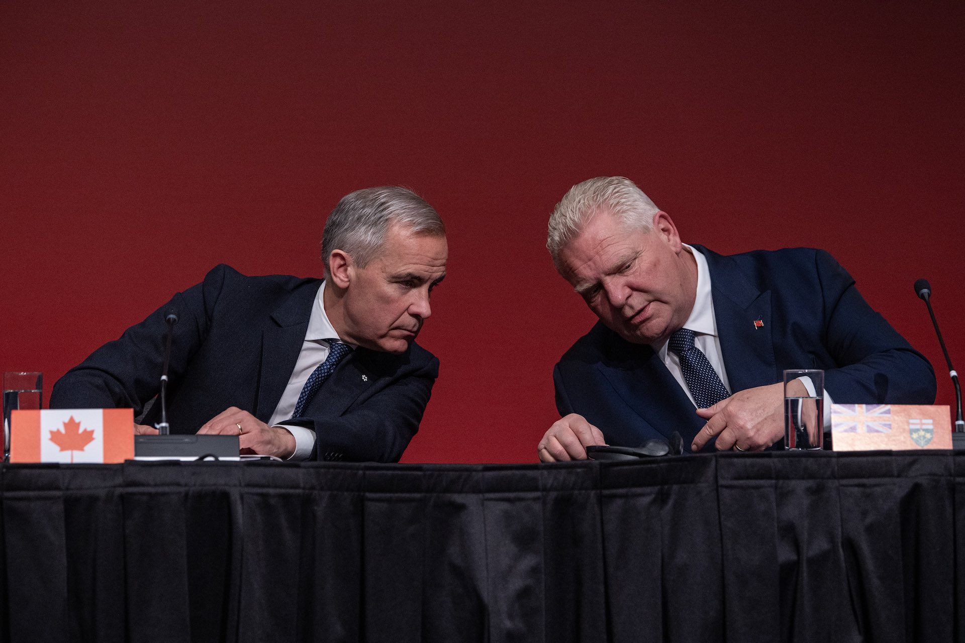A shot of Mark Carney and Doug Ford at a press conference table, leaning their heads toward each other as they confer privately. There are microphones on the table, and a red backdrop behind them.