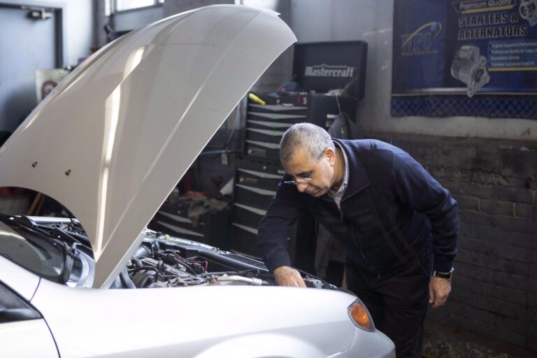 A mechanic in an auto repair shop looks under the hood of a silver car.
