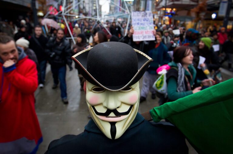 A close-up of a man wearing a Guy Fawkes mask, a black jacket and a black hat in the midst of a large protest parade. The mask appears to be mounted on the back of the person's head, not over his face.
