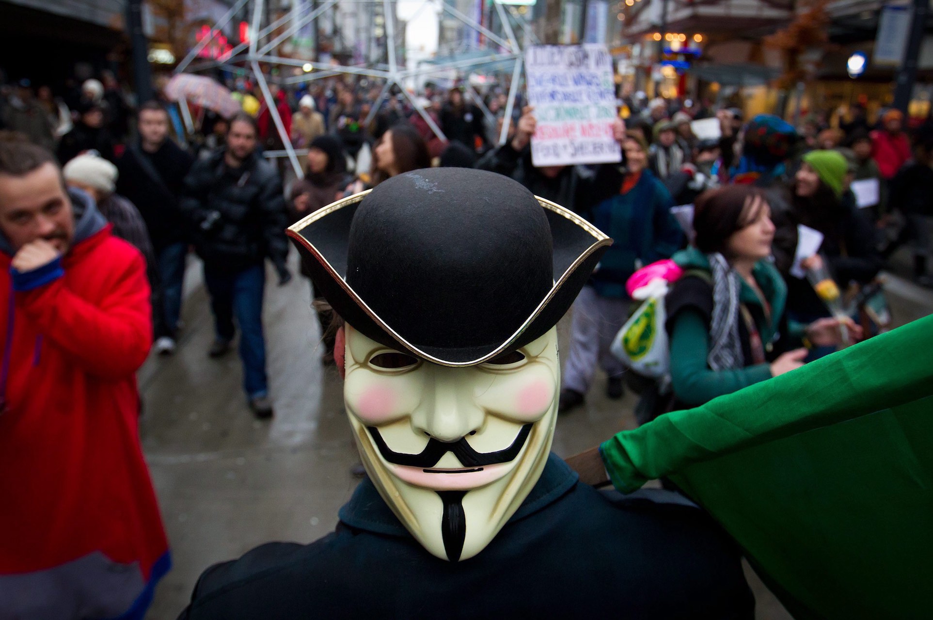 A close-up of a man wearing a Guy Fawkes mask, a black jacket and a black hat in the midst of a large protest parade. The mask appears to be mounted on the back of the person's head, not over his face.