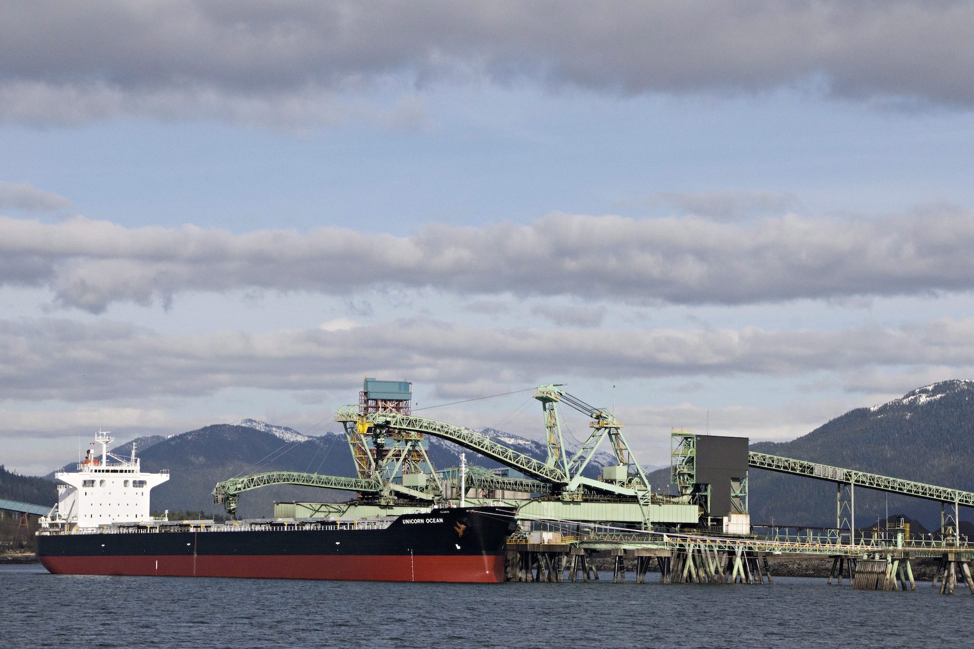 A wide shot of large ship at a dock; it's being loaded from metal booms bearing conveyors. There are snow-capped mountains in the background.