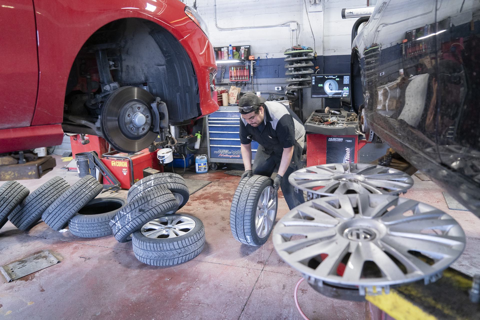 A mechanic rolls a tire toward a red car on a hoist in an auto shop. The car's front brake assembly is visible in the top left corner of the frame.