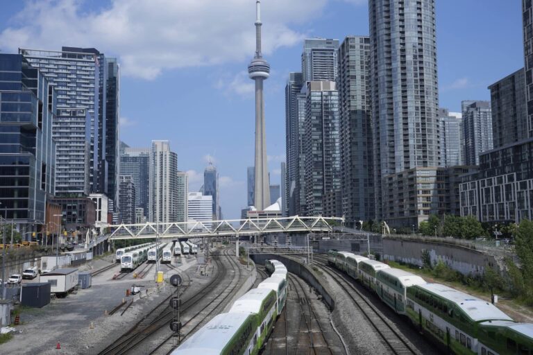 View of multiple train tracks and green trains in downtown Toronto, with high-rise buildings and the CN Tower in the background under a partly cloudy sky.