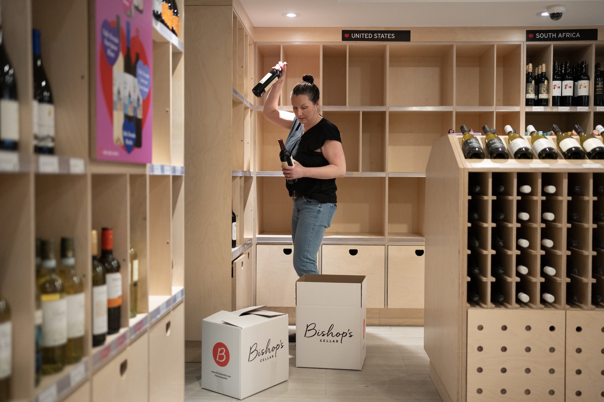 A shot of a liquor-store employee with a wine bottle in each hand and two white, cardboard boxes at her feet. There is an empty floor-to-ceiling set of shelves behind her labelled "United States."
