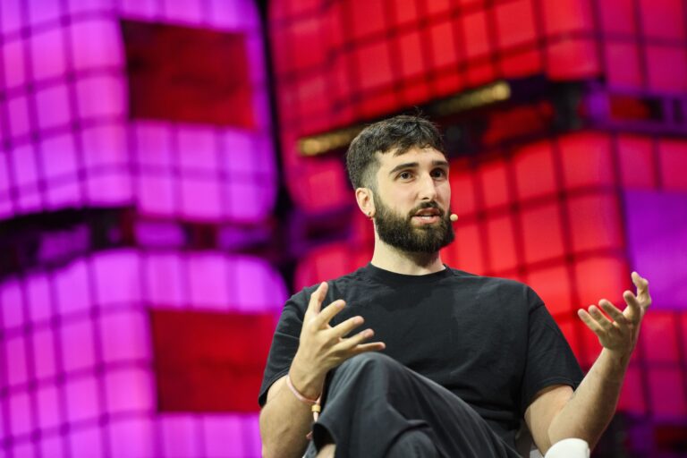 A man in a black T-shirt sits on a stage gesturing with both hands as he speaks. Behind him is a lit red-and-purple stage backdrop