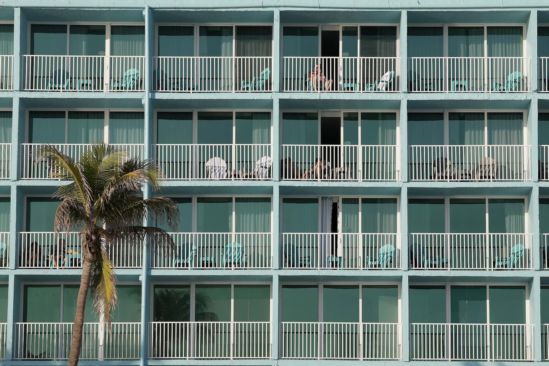 Four stories of apartment balconies fill the frame. A single palm tree juts into frame in the lower left. Two balconies have a person relaxing, barely visible through white railings