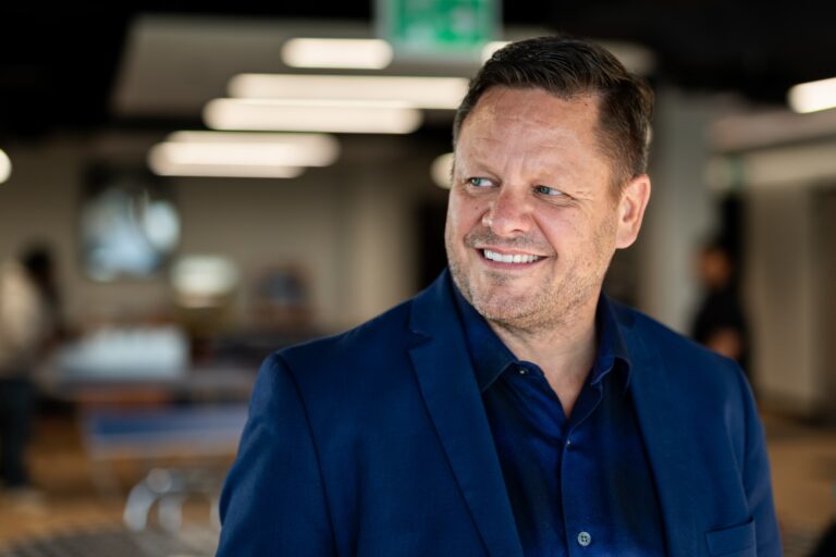 A man in a blue shirt with blue blazer smiles as he looks slightly back over his shoulder. The background is blurred but shows an office setting.