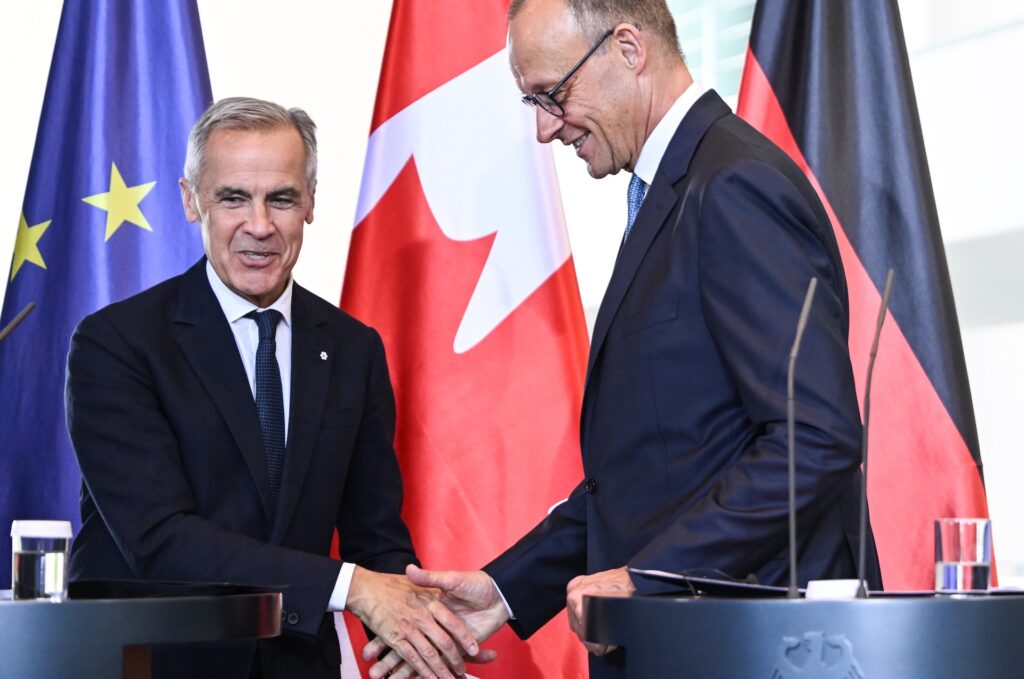 Mark Carney shaking hands with German Chancellor Friedrich Merz, with EU, Canadian and German flags in the background. Both men are smiling.