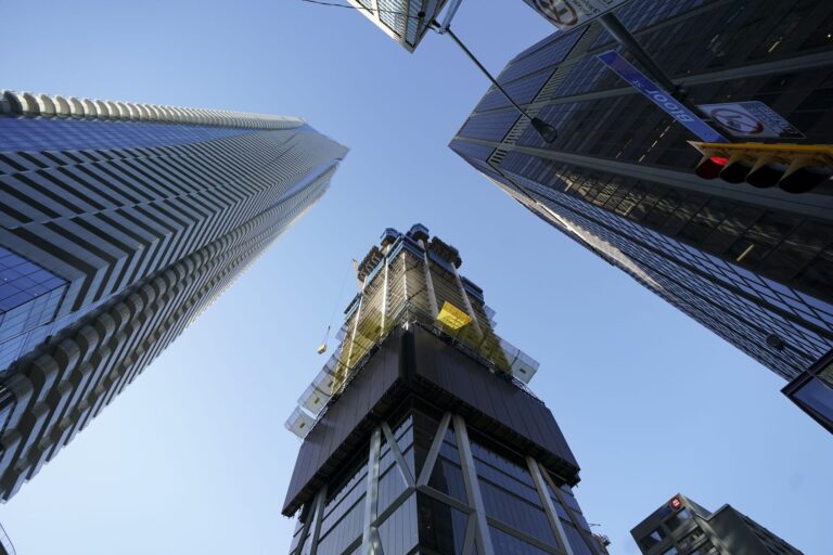 A view of of several tall skyscrapers, including one under construction, against a clear blue sky in an urban cityscape.