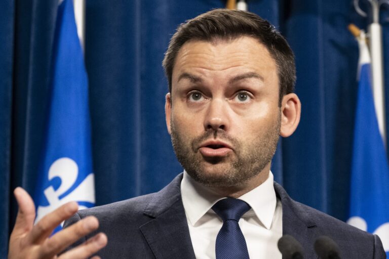 A man gestures with an open hand while speaking in front of blue curtains and a pair of Quebec flags.