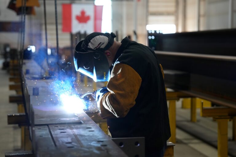 A close-up of a worker welding in an industrial plant; there is a Canadian flag on the wall in the background.