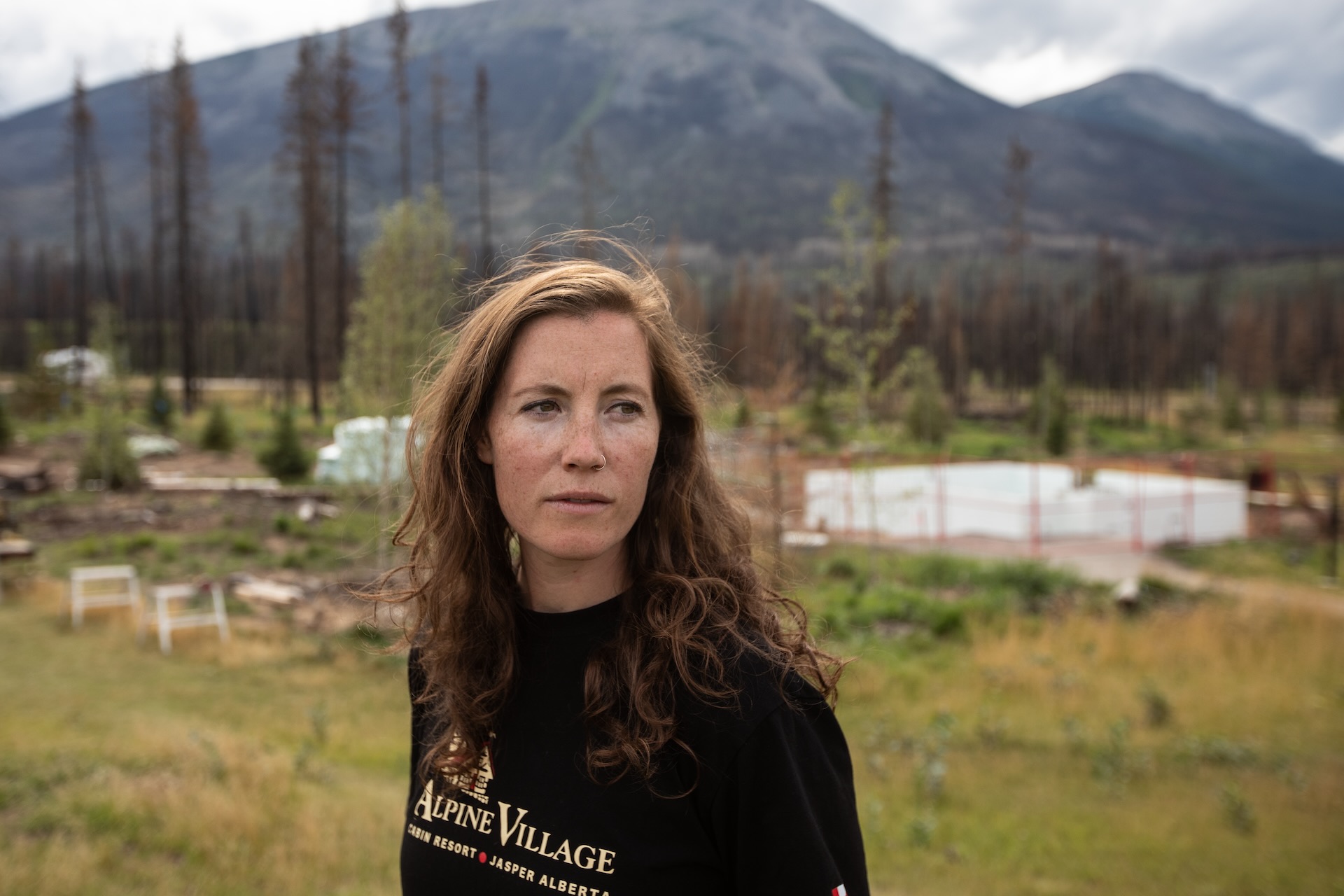 A woman in a black sweater with a logo for Alpine Village Cabin Resort in Jasper, Alta. looks to her left in the foreground. In the distance behind her is a blur of trees and mountains.