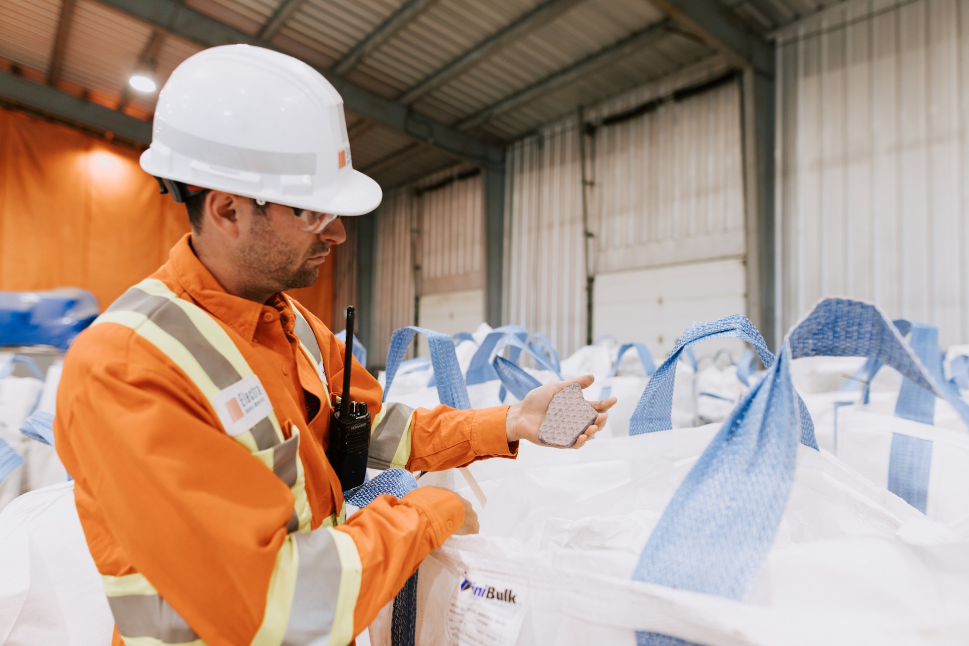 A worker wearing a white hardhat and an orange work shirt holds a piece of mineral product in his left hand. He's in an industrial building filled with large bags.