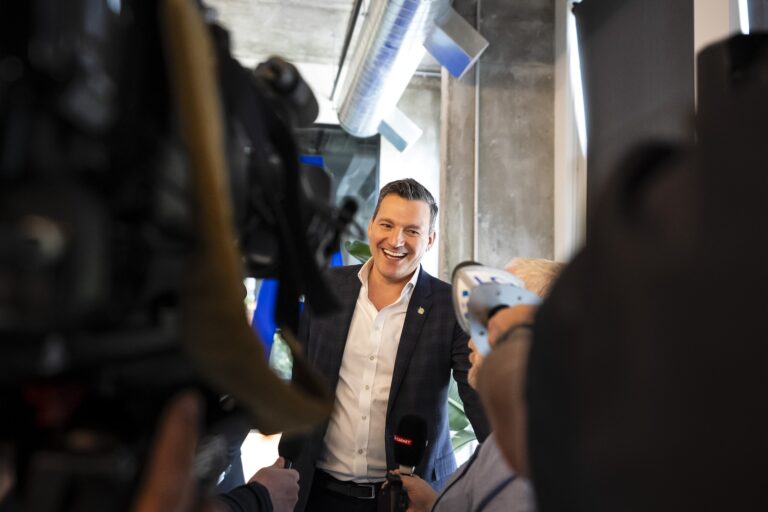 Evan Solomon, in a white collar shirt and blazer with no tie, smiles as he speaks to reporters. The picture is taken from behind some reporters with camera equipment and shoulders in the foreground, framing Solomon.