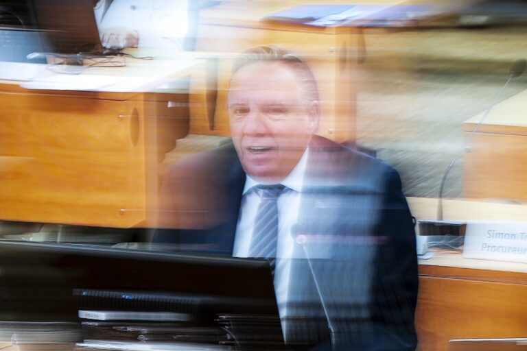 A man in a suit speaking into a microphone at a desk with a computer screen facing him. There is a white blur effect over much of the image.