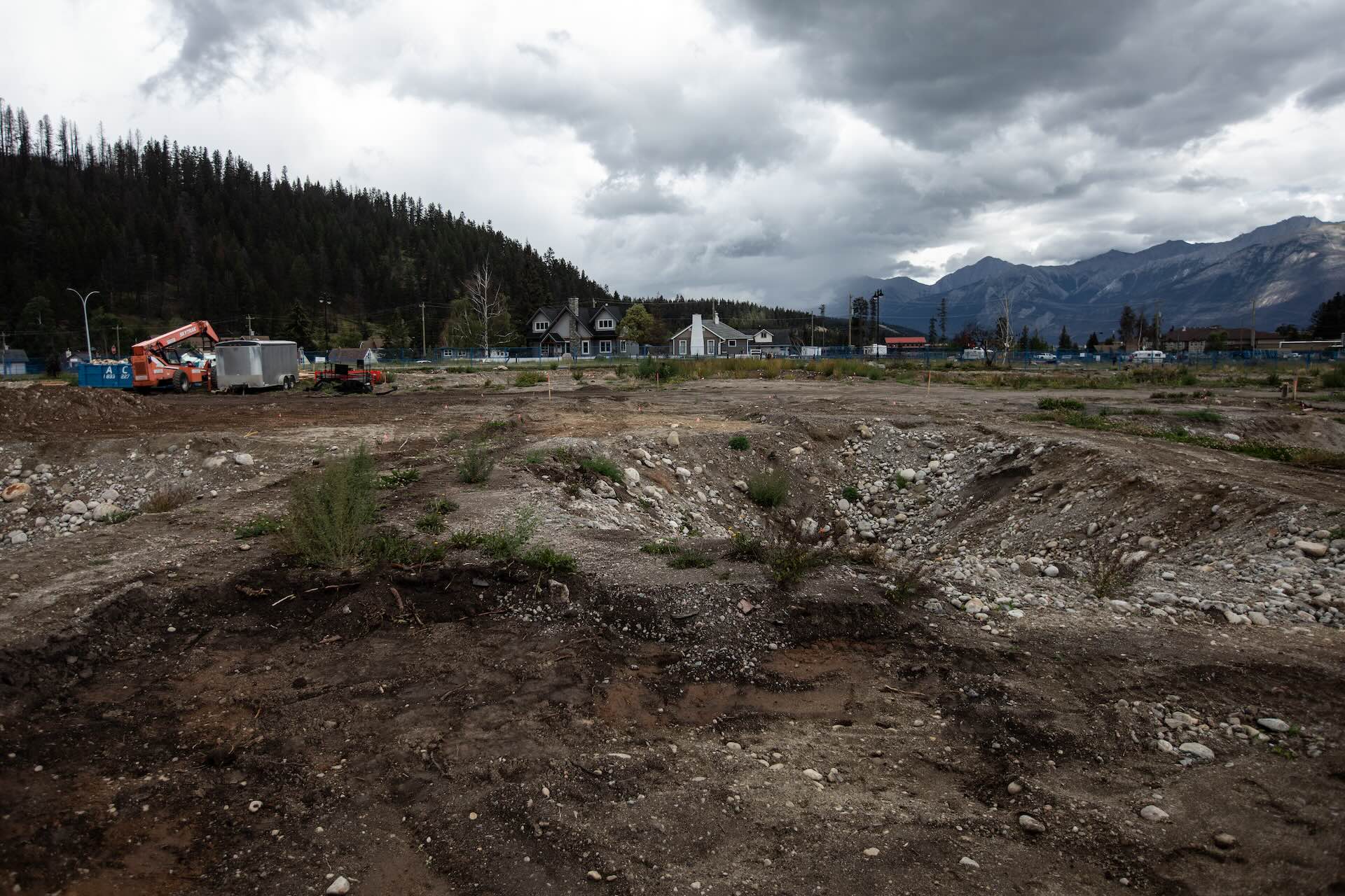 A divot in a dirt field is seen in the foreground. The middle distance shows utility trucks and a handful of houses, with tree-lined hills and mountains under a grey sky in the background.