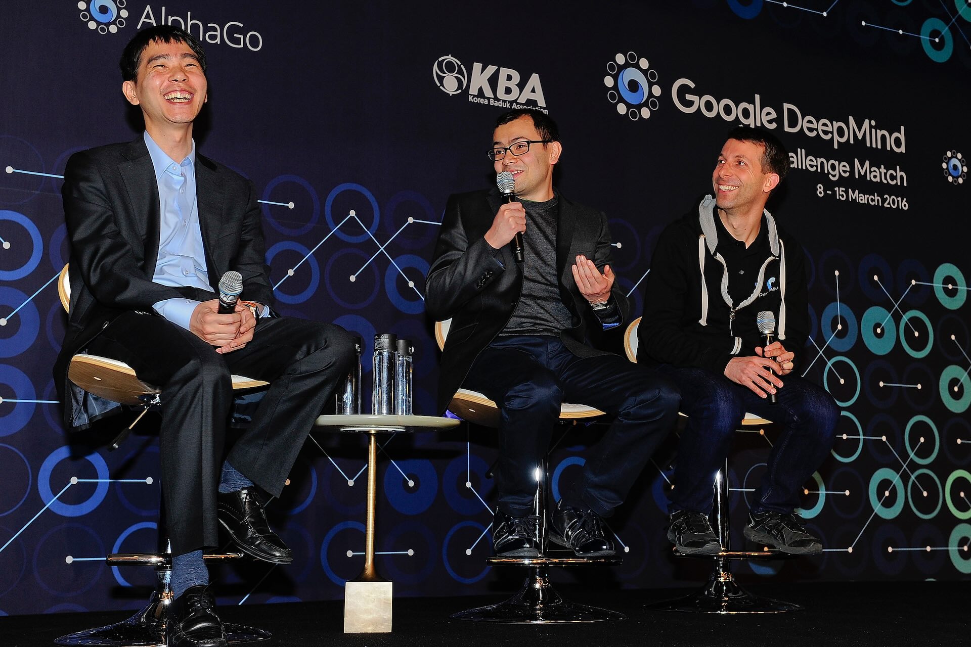 Three men sitting on a stage in business casual dress against a backdrop with logos for Korea Baduk Association and Google DeepMind. The man in the middle is speaking into the microphone with a smile on his face, and the men on either side of him are smiling broadly