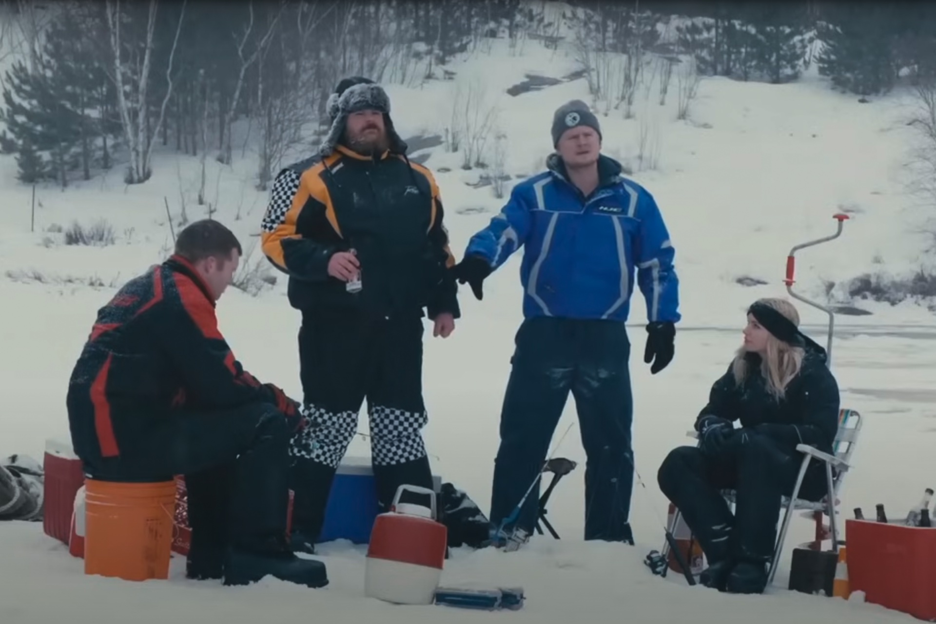 A shot from the 'Les hiques' scene in Letterkenny, with four characters wearing snowmobile suits and drinking beer while ice fishing. Two are standing and appear angry.