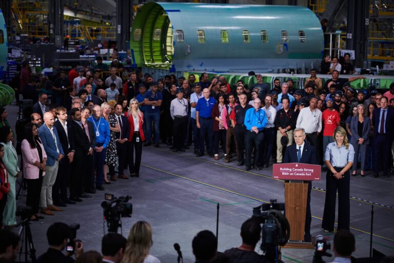 A high-angle wide shot of Mark Carney at a lectern, with Mélanie Joly standing to his left and a crowd of people behind him. They are in a cavernous, hangar-like building and there is a large segment of airplane fuselage in the distant background.